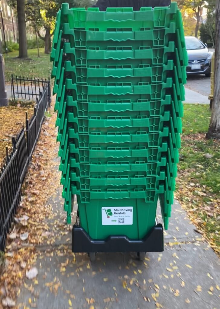 Stack of green reusable moving boxes on a delivery dolly from “Mai Moving Rentals” on a Toronto sidewalk.